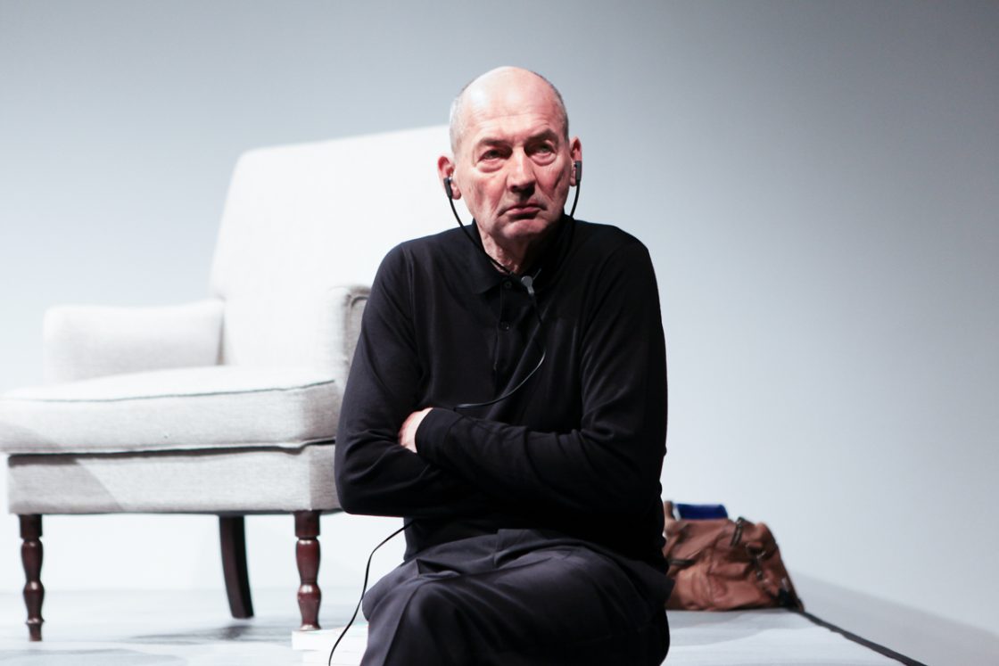 Rem Koolhaas sentado no palco, vestindo roupa preta, usando fones de ouvido e olhando fixamente para a frente, com uma poltrona clara e uma bolsa marrom ao fundo