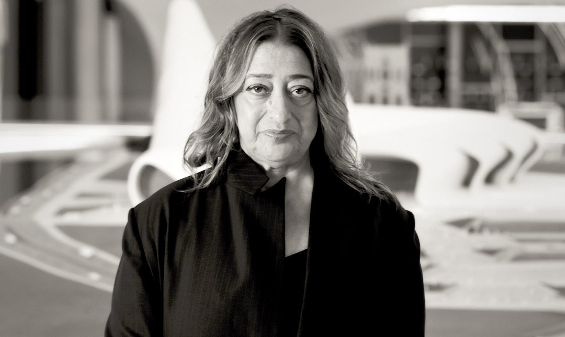 Fotografia em preto e branco de Zaha Hadid em pé diante de uma maquete arquitetônica de linhas curvas; ela usa um casaco escuro e olha para a câmera com expressão serena