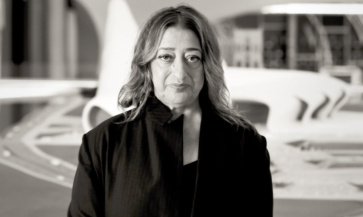 Fotografia em preto e branco de Zaha Hadid em pé diante de uma maquete arquitetônica de linhas curvas; ela usa um casaco escuro e olha para a câmera com expressão serena