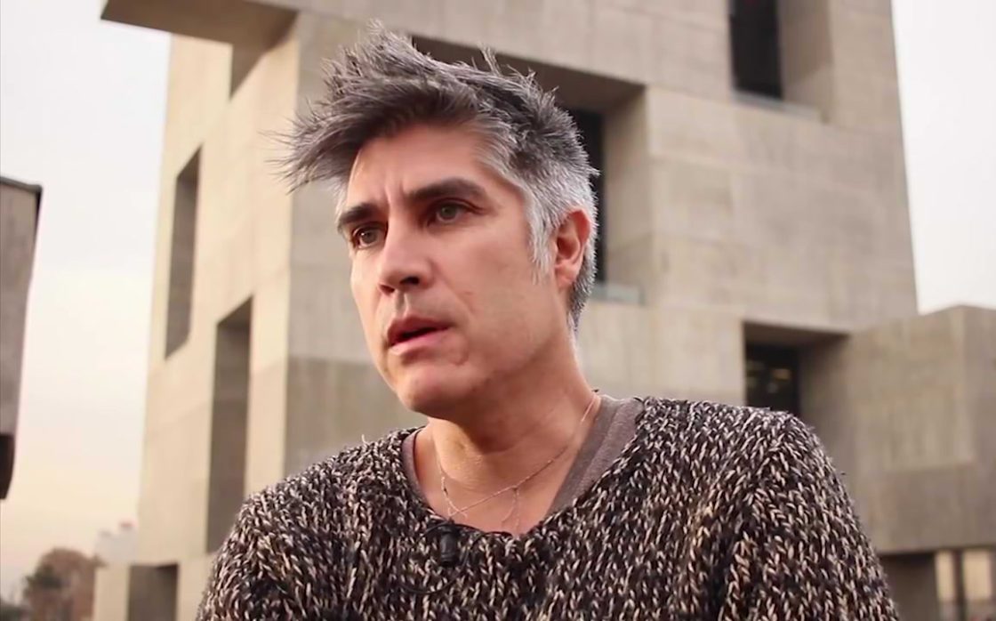 Alejandro Aravena em primeiro plano, usando suéter escuro, com edifício de concreto ao fundo e expressão concentrada