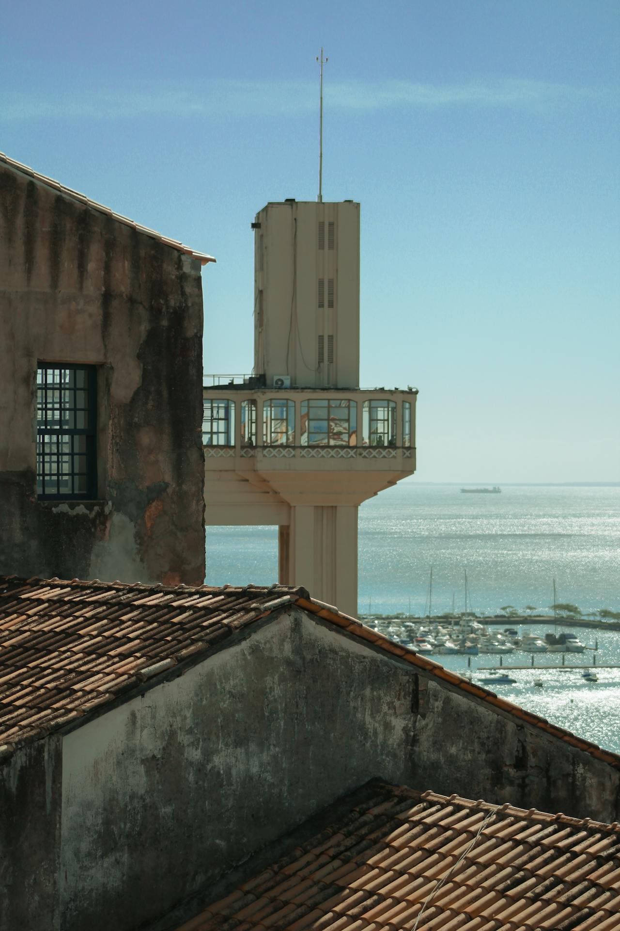 Estrutura do Elevador Lacerda vista ao lado de telhados históricos da Cidade Alta, evidenciando a torre em concreto e a plataforma suspensa com vista para o mar de Salvador