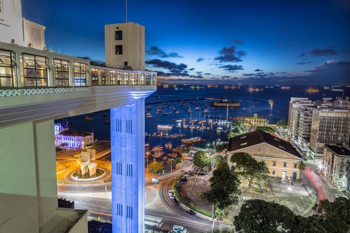 Vista noturna do Elevador Lacerda, em Salvador, com iluminação cênica, barcos na baía, Mercado Modelo e vias urbanas movimentadas ao redor da estrutura