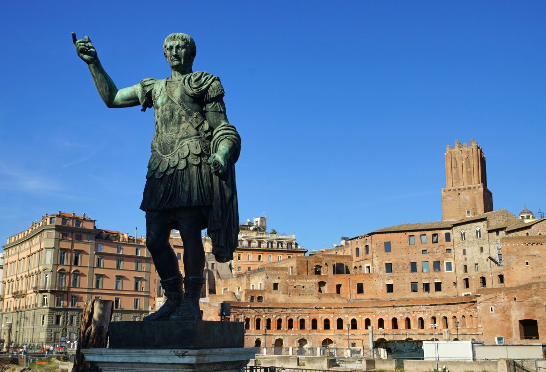 Estátua do Imperador Trajano na Rua dos Fóruns Imperiais em Roma, com casas do Mercado de Trajano ao fundo.