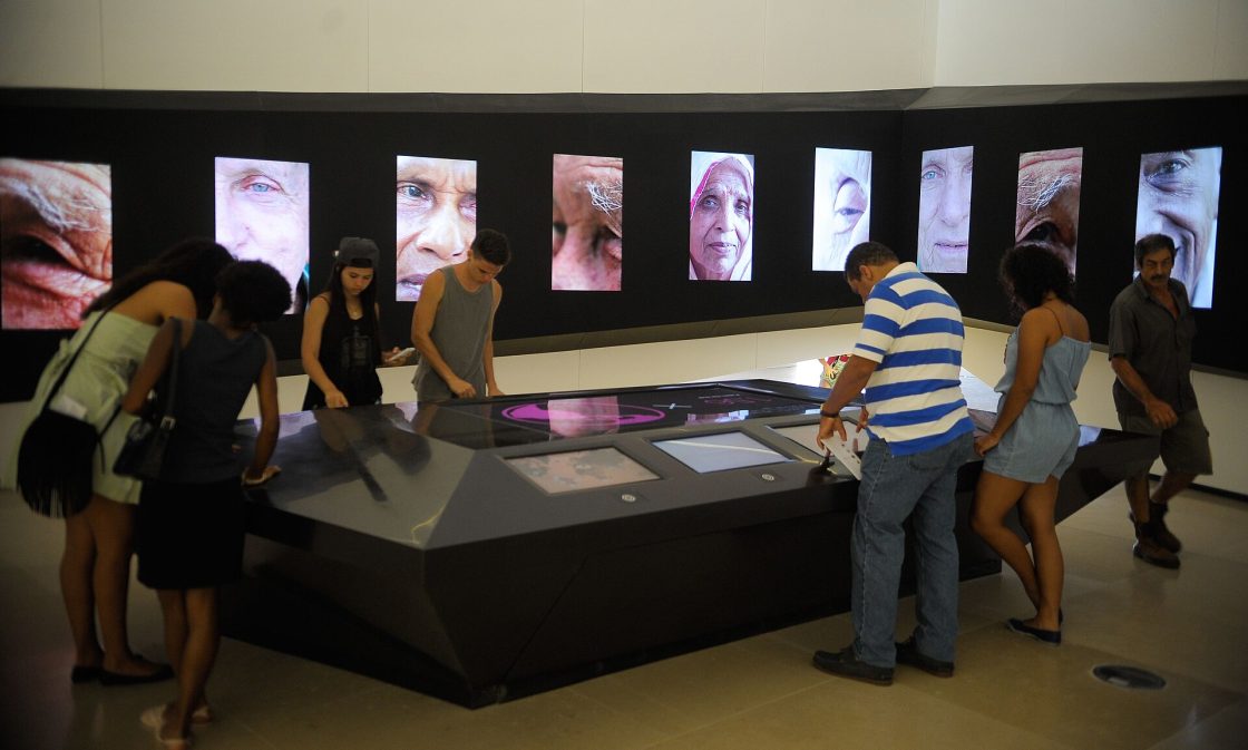 Pessoas no entorno de mesa interativa que faz parte de exposição permanente do Museu do Amanhã