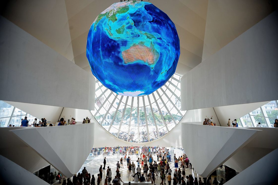 Visão de dentro para fora do Museu do Amanhã, com muita luz natural entrando e grande globo terrestre preso ao teto, que tem pé-direito alto