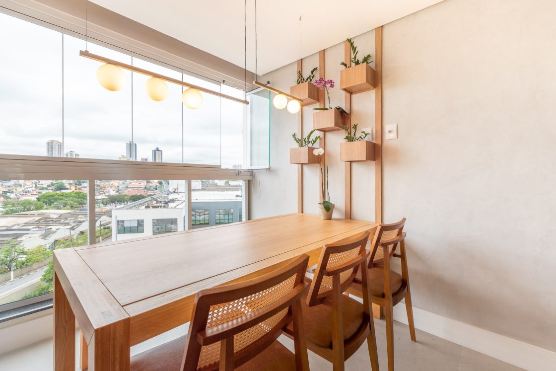 Varanda de apartamento com cimento queimado na parede. Ela também conta com mesa, cadeiras, lustre e vasos