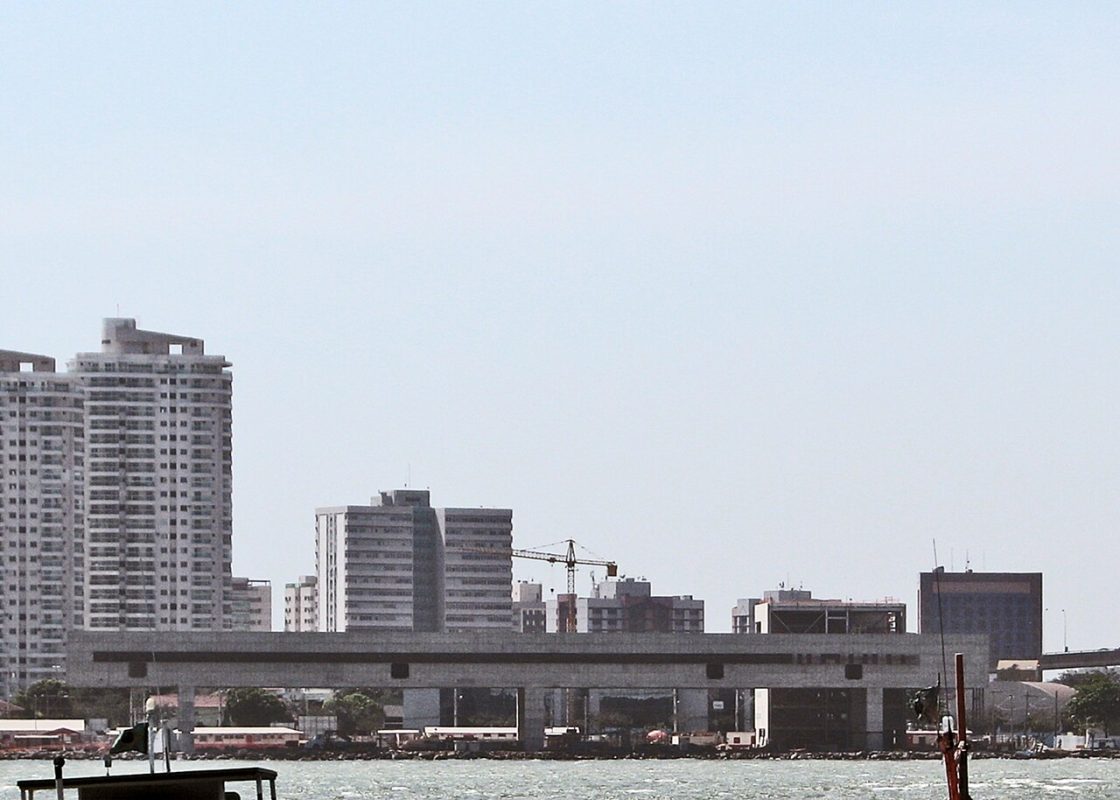 Imagem do Cais das Artes mostrando edifício alongado em concreto apoiado em pilares, com skyline urbano e guindaste ao fundo diante da orla de Vitória
