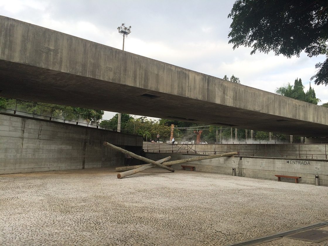 Vista externa do MUBE mostrando cobertura horizontal de concreto sustentada por pilares, área rebaixada com bancos e obra escultórica ao centro