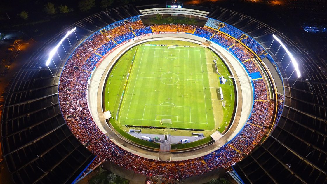 Vista aérea noturna do Estádio Serra Dourada lotado, com arquibancadas circulares iluminadas e campo de futebol ao centro