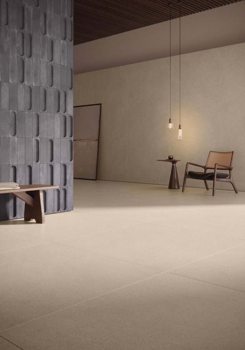 Detalhe de ambiente com piso bege e parede 3D cinza. Do lado direito, há uma cadeira, uma mesinha e uma lâmpda pendente