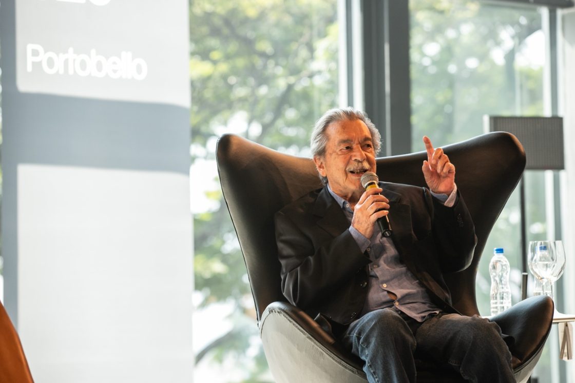 Paulo Mendes da Rocha sentado em poltrona preta, segurando microfone e apontando com o dedo durante palestra no Archtrends Summit, com painel da Portobello ao fundo