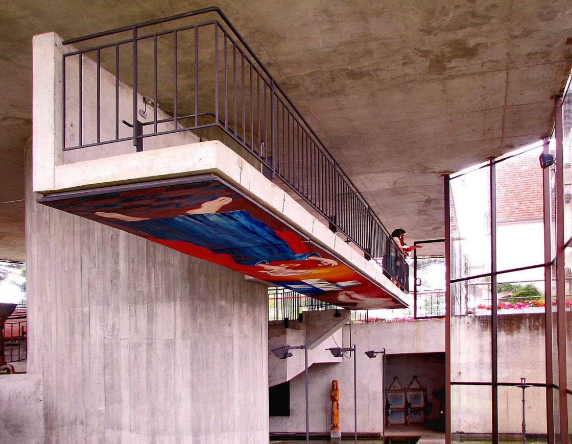 Vista interna da Capela de São Pedro mostrando volumes de concreto, escada geométrica, grandes planos envidraçados e uma pessoa apoiada no guarda-corpo superior