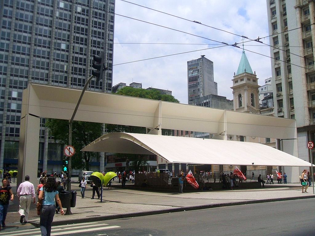 Pórtico da Praça do Patriarca em São Paulo com estrutura metálica branca e cobertura curva, cercado por prédios altos e pedestres atravessando a praça
