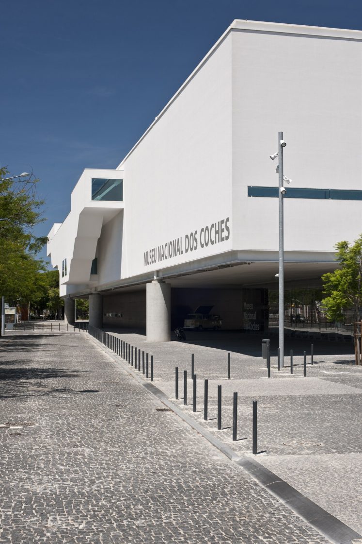 Vista externa do Museu Nacional dos Coches mostrando bloco branco elevado sobre pilotis, entrada envidraçada e passeio público com balizadores metálicos