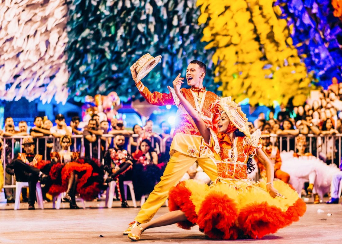 Apresentação de quadrilha junina com dançarinos em roupas tradicionais coloridas e público acompanhando o espetáculo em evento festivo noturno