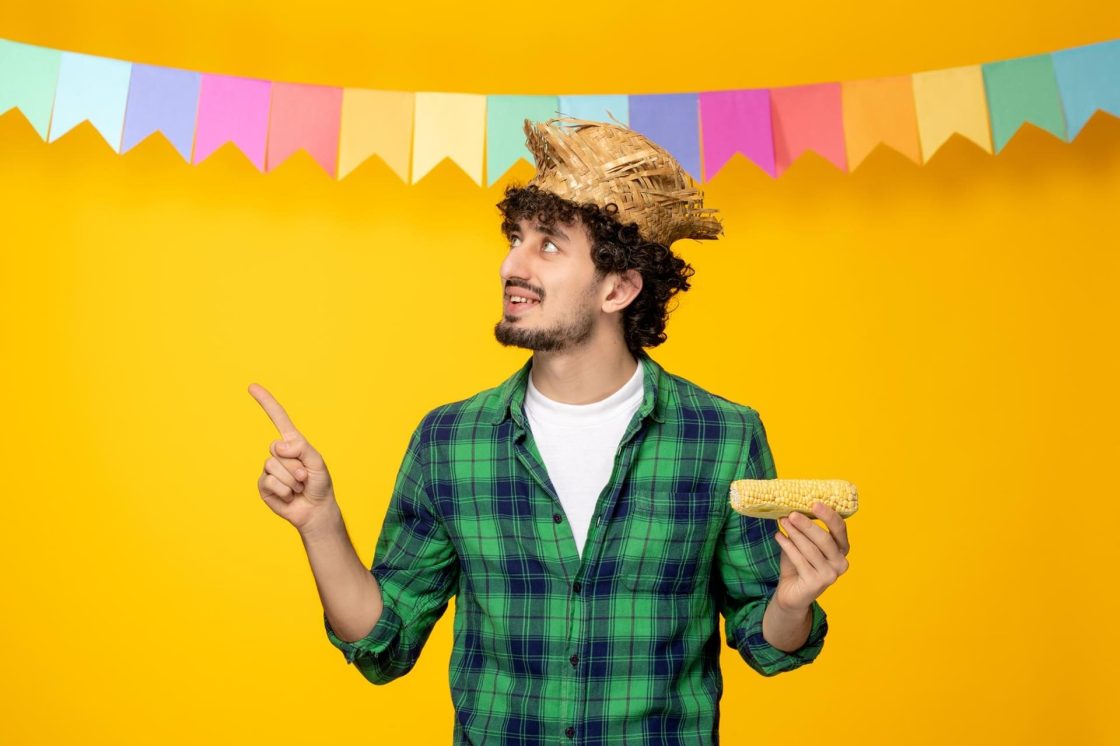 Homem com camisa xadrez, chapéu de palha e espiga de milho posando diante de fundo colorido com bandeirinhas em cenário temático de festa junina