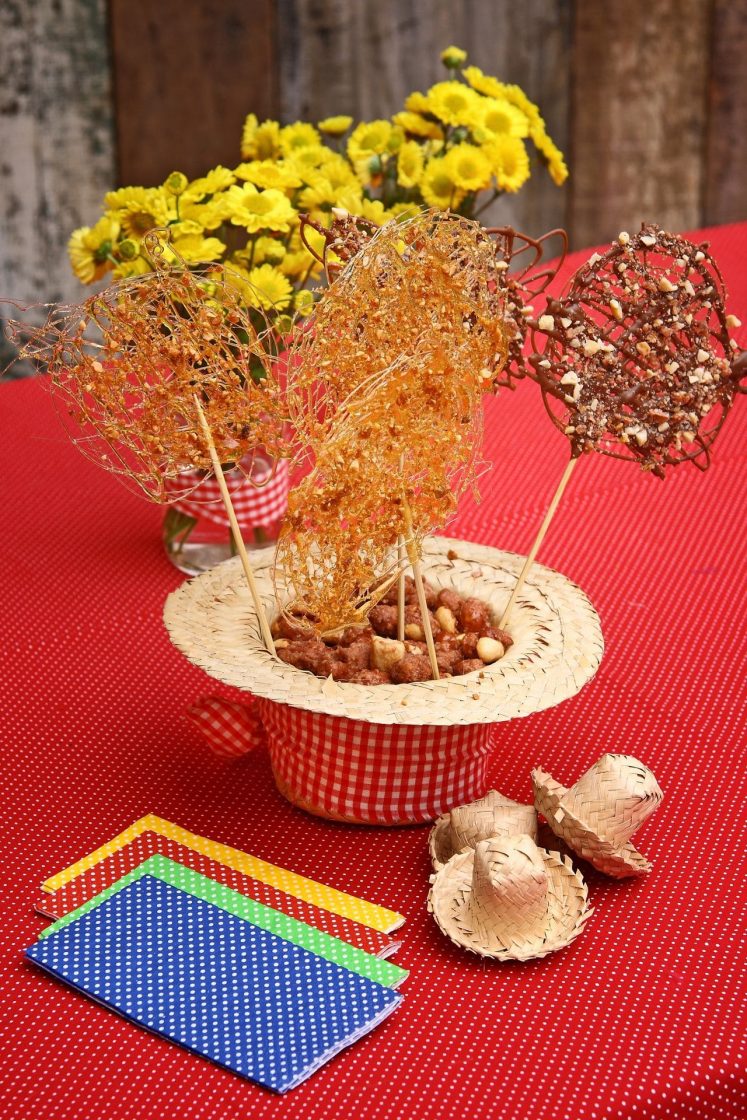 Mesa de festa junina com tecido xadrez vermelho, chapéu de palha decorativo, doces típicos e flores amarelas ao fundo