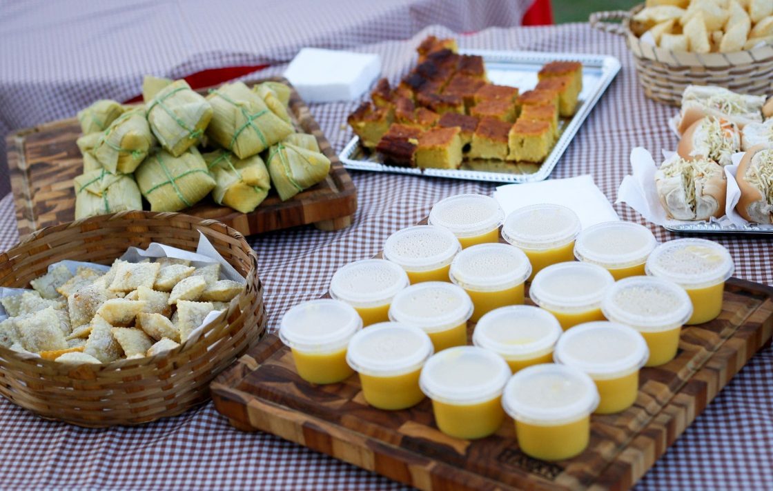 Mesa de quitutes juninos com canjica, pamonha, bolo de milho, doces típicos e toalha xadrez, valorizando a gastronomia como elemento decorativo