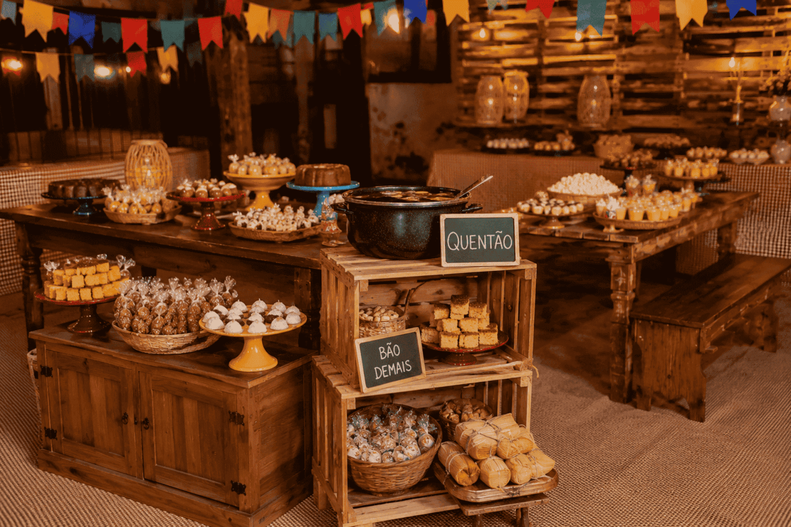 Ambiente de festa junina com mesas e bancos de madeira aparentes, caixotes reutilizados como expositores de quitutes típicos, panela de quentão e bandeirinhas coloridas compondo uma decoração rústica e acolhedora