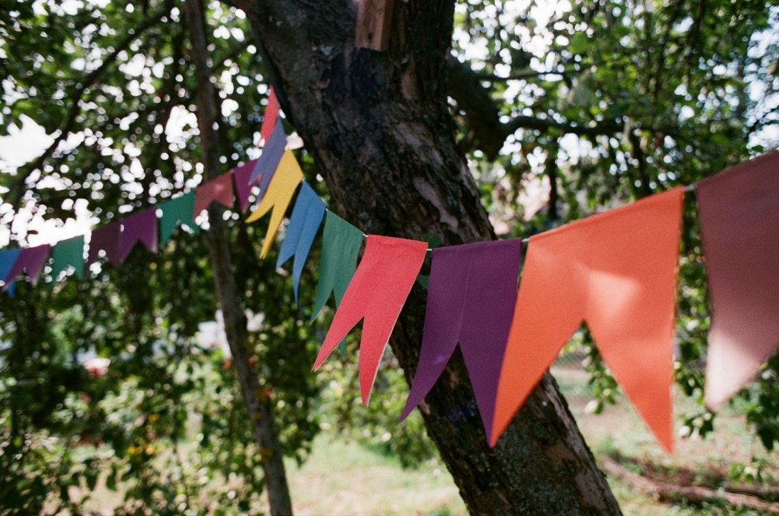 Cordão de bandeirinhas coloridas pendurado entre árvores em área externa, criando uma ambientação típica de festa junina