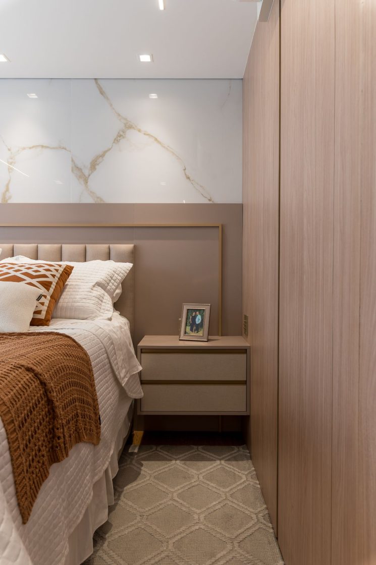 Cama de casal com roupa de cama branca e em tom terroso, com mesa de cabeceira com foto de casal ao lado