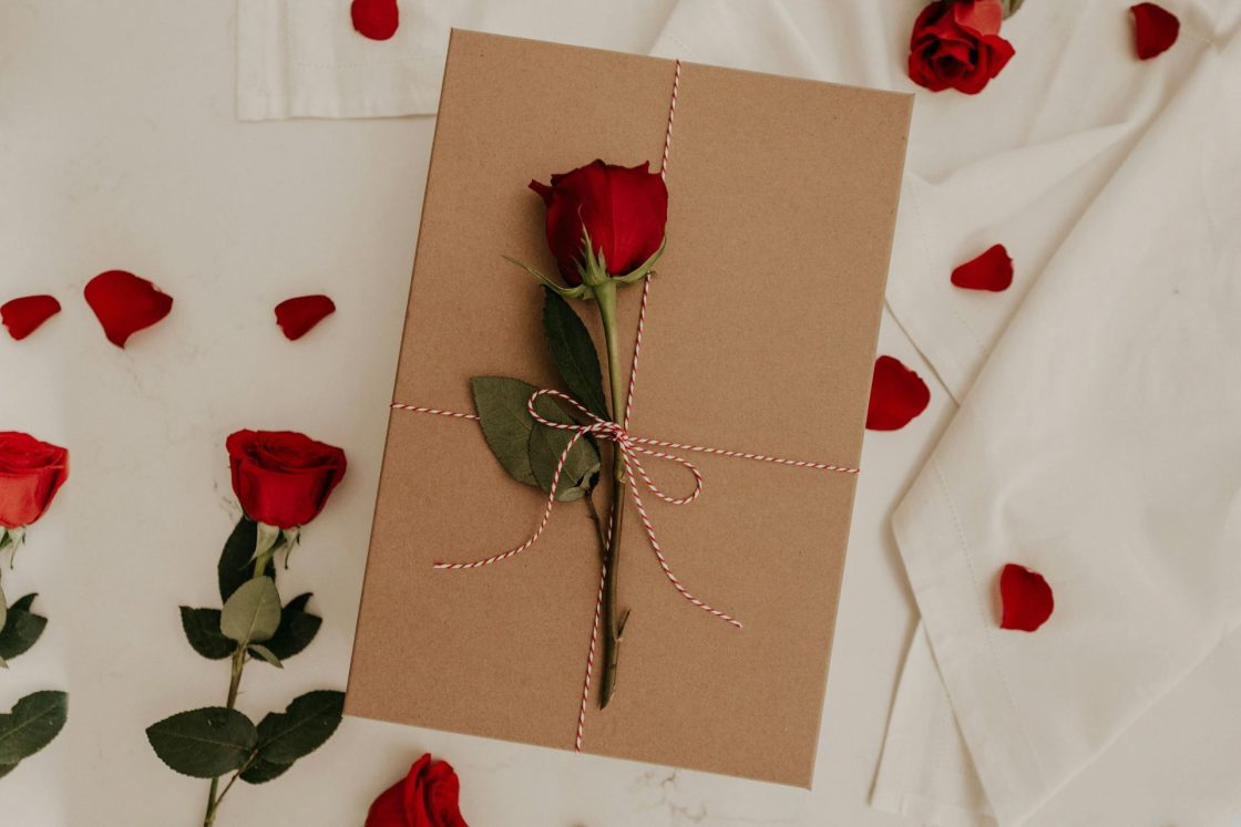 Cartão em papel craft com rosa vermelha por cima, amarrada com fita branca e vermelha