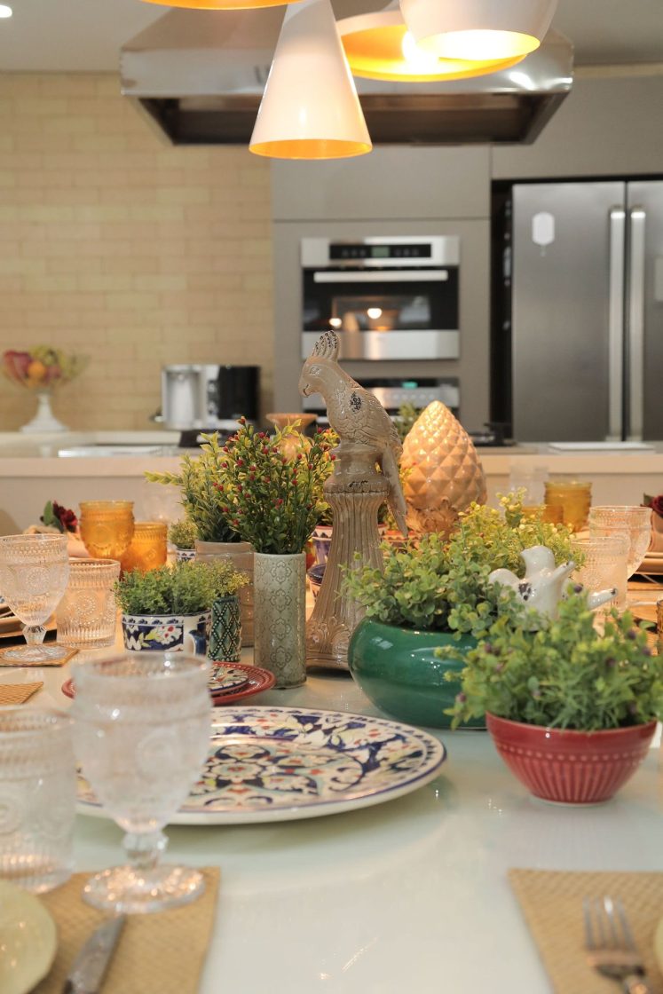 Mesa posta com pratos decorados, taças transparentes e diferentes arranjos com plantas verdes