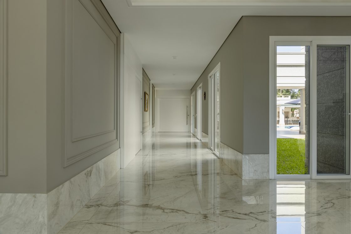 Corredor interno com piso de porcelanato, painéis de parede estilo boiserie e uma grande porta de vidro que se abre para um quintal gramado com piscina