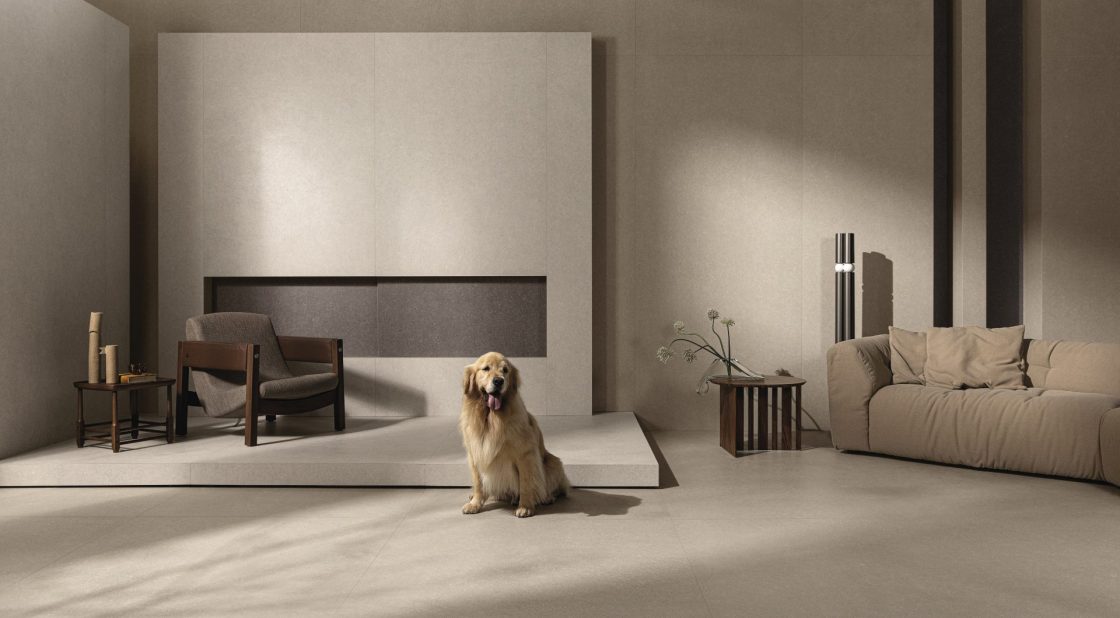 Living com revestimento que reproduz limestone em tom claro, sofá bege, poltrona de madeira estofada em cinza e cachorro da raça golden retriever