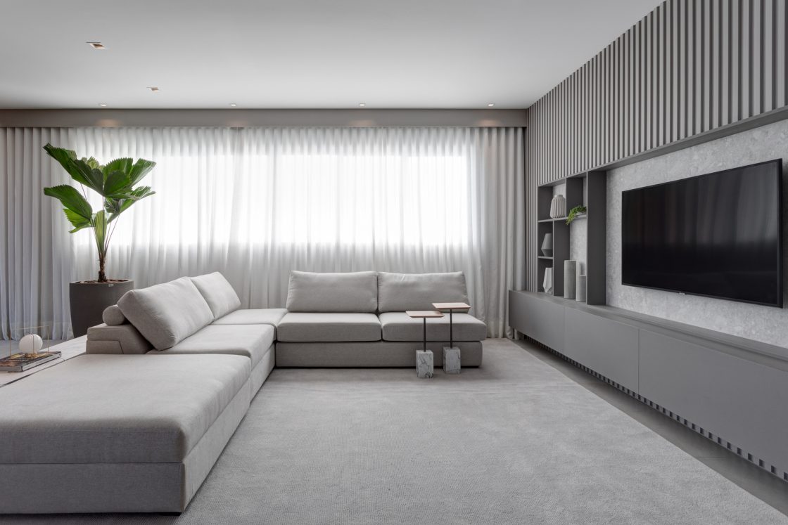 Sala de TV com tons claros de cinza no sofá, tapete e ripado de madeira, ambiente com muita luz natural