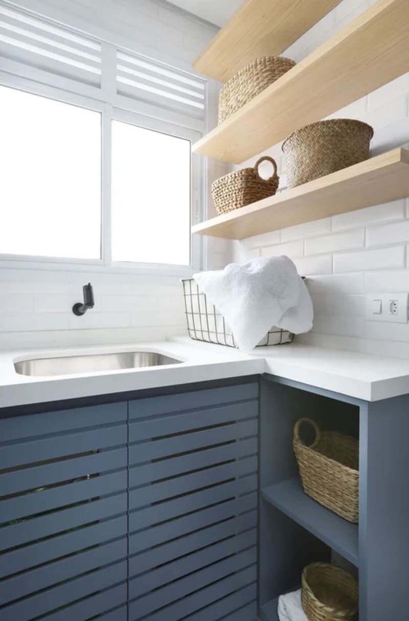 área de lavar com pia clara branca com uma torneira, cesto de roupas, e três prateleiras suspensas em madeira