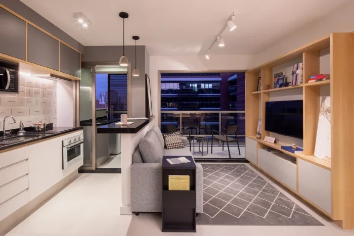Cozinha americana com fogão embutido, armário branco, divisória de parede com sofá cinza, televisão e painel para televisão, tapete cinza geométrico no chão 
