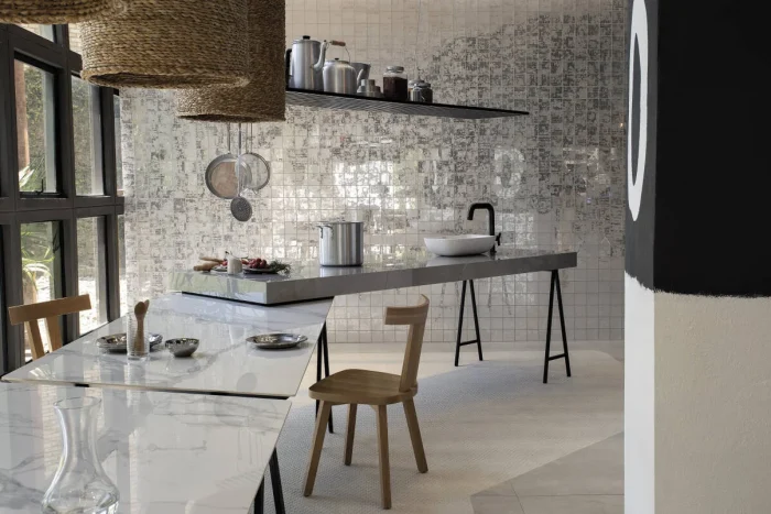 Cozinha com bancada minimalista e moderna integrada a uma mesa mais baixa e com cadeiras de madeira. A parede é revestida por pastilhas em tons de cinza.