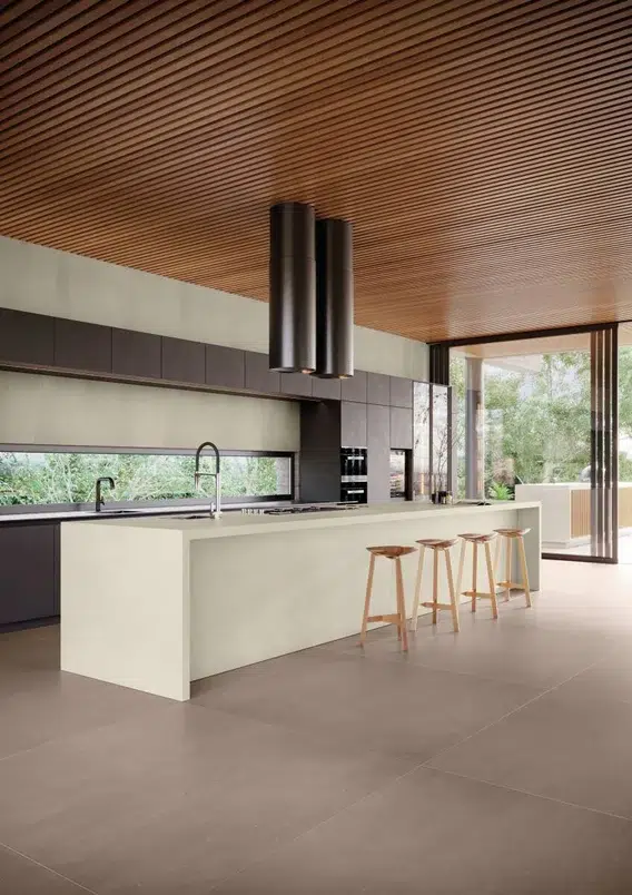 Cozinha com longa bancada clara e quatro banquetas de madeira. Móveis escuros e decoração minimalista em um ambiente com piso que reproduz a aparência de concreto.