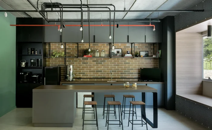 Cozinha moderna com móveis escuros e parede de tijolinhos aparentes. O projeto é no estilo industrial, por isso é possível bastante ferragens nos elementos e pendentes simples para iluminação.
