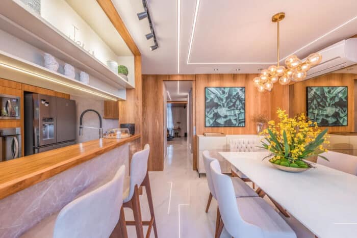 Cozinha integrada com sala de jantar, revestimento na parede e bancada toda de madeira, mesas e cadeiras brancas e eletrodomésticos nas tonalidades cinza 