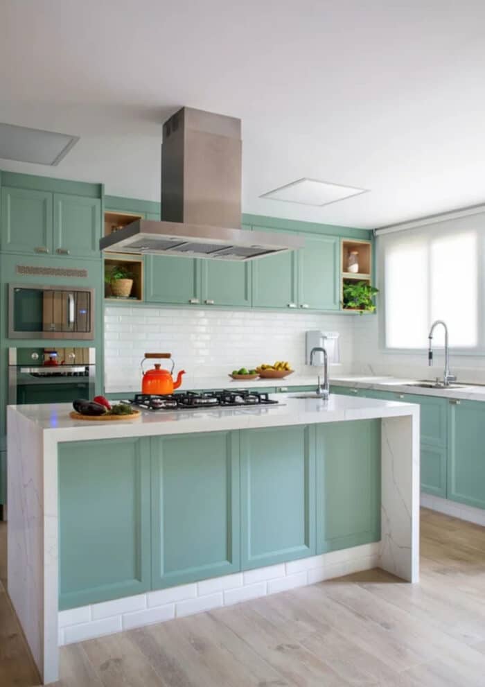 Cozinha com ilha pequena central em tons de verde, bancada em mármore branco e chaleira laranja em cima do cooktop 
