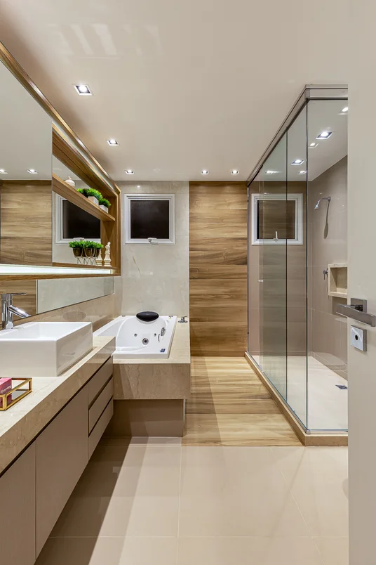 Banheiro amplo com iluminação embutida no teto, box de vidro e banheira retangular ao lado. Armário embutido com pia única quadrada 