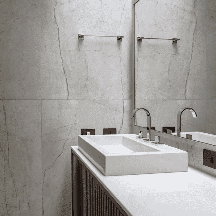 Banheiro com parede toda em mármore na tonalidade cinza. Pia branca quadrada e armário em madeira