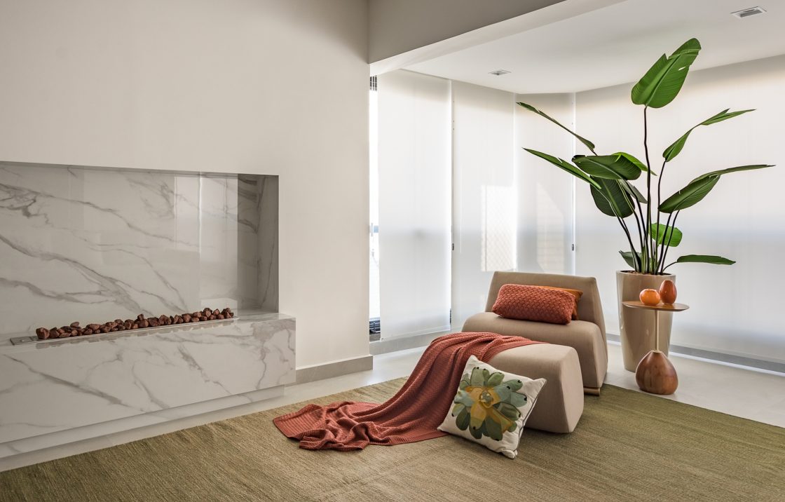 Lareira em porcelanato com poltrona com manta e almofadas em tons terrosos em frente