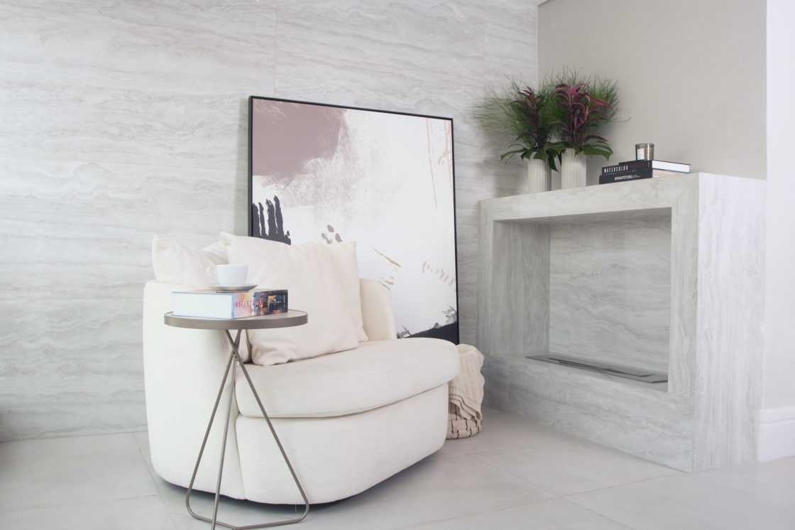 Ambiente em tons claros com poltrone em frente à lareira com porcelanato marmorizado que reproduz travertino