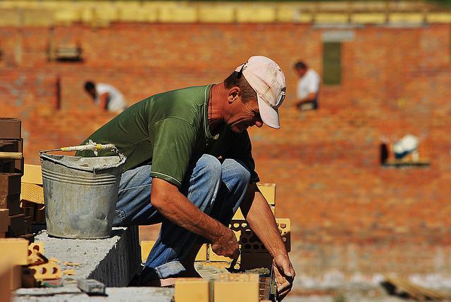 Pedreiro com boné branco e blusa verde colocando os tijolos em uma construção.