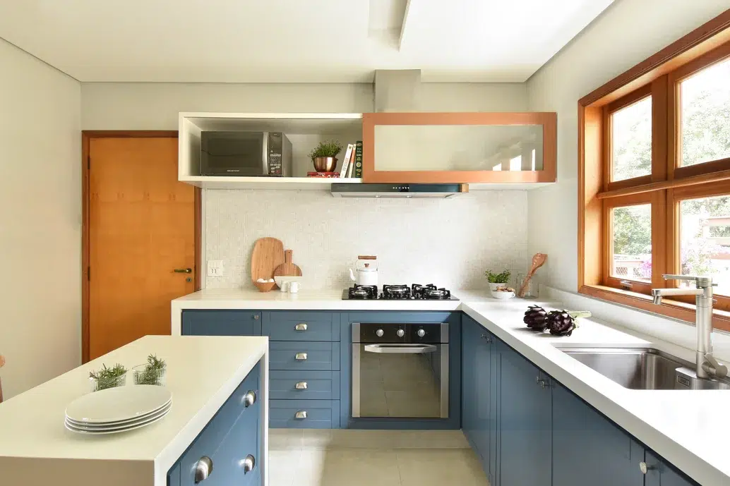 Cozinha com móveis em azul petróleo e a mesa em cor branco. Uma janela com marco de madeira.