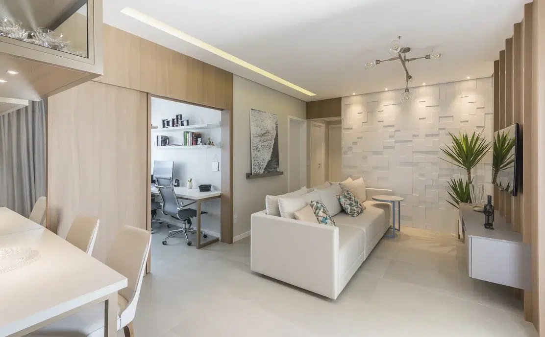 Sala de apartamento pequeno com uma parede em 3d de retângulos pequenos, sofá off white com almofada de cores, tv na parede 3d de madeira. 