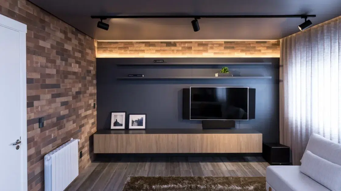 Sala de estar de um apartamento com uma parede de tijolos e a outra parede preta com uma tv, um rack com dois quadros, chão de madeira e cortina comprida.