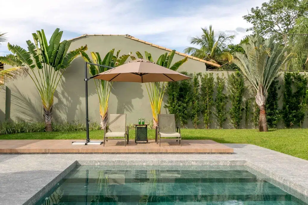 área externa de uma casa, jardim com piscina e dois cadeiras de praia embaixo de um guarda-sol.