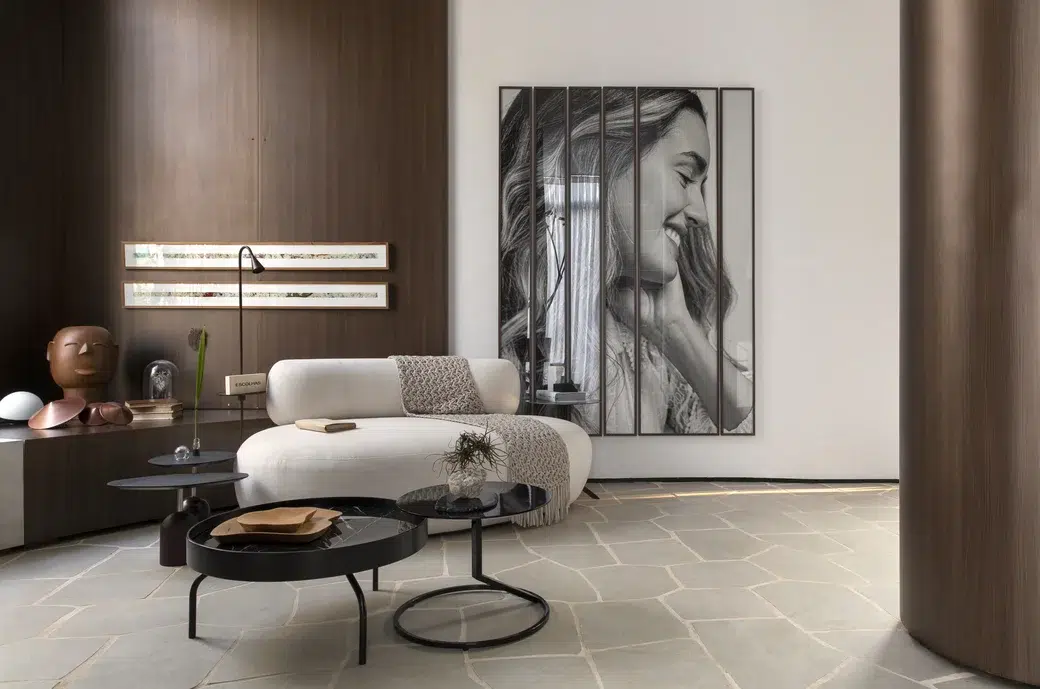 sala de estar de apartamento com um sofá pequeno off white, parede com revestimento de madeira marrom escura, quadro gigante de uma mulher sorrindo de perfil em preto e branco. Duas mesas pequenas de centro pretas.