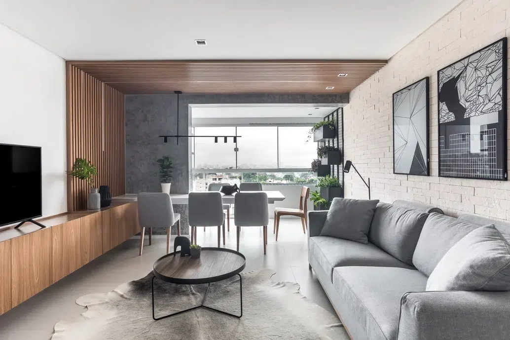 Sala de estar e mesa de jantar no mesmo ambiente de um apartamento, estilo industrial com decoração em cores cinza e preto. Metade do teto com revestimento de madeira e quadros  em cores preto e branco em uma parede de tijolos.