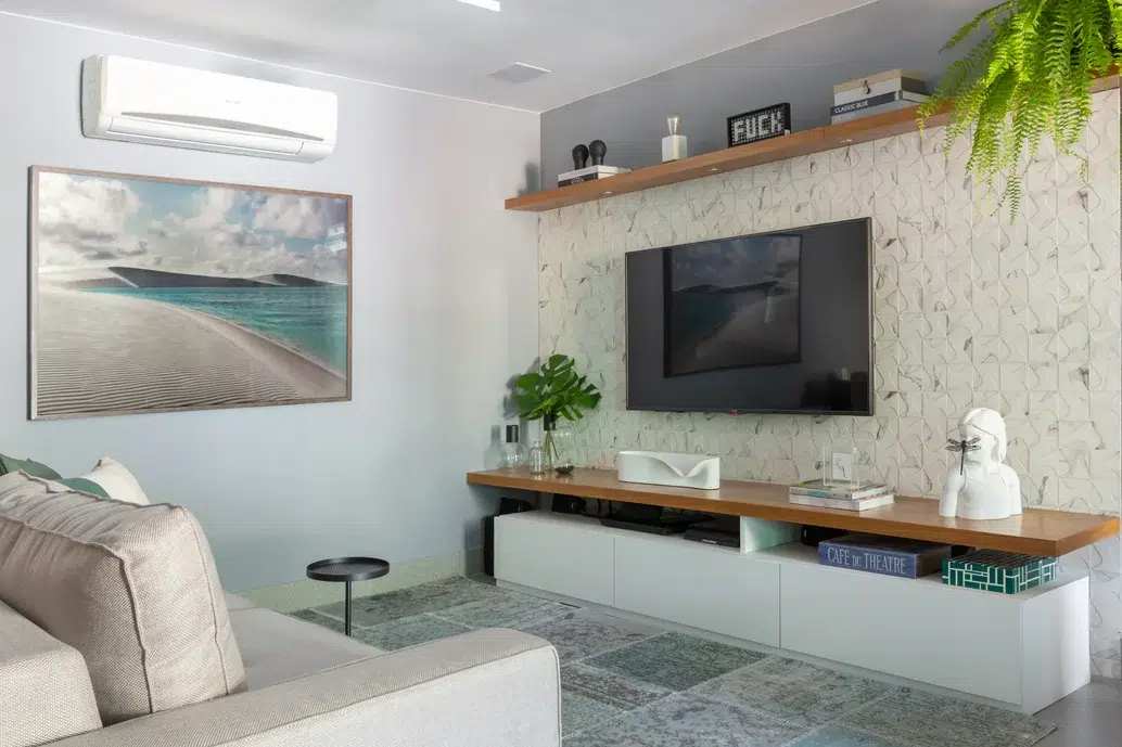 Sala de estar de um apartamento com uma parede texturizada com a tv pendurada, decoração de plantas e livros, um quadro de paisagem e sofá.