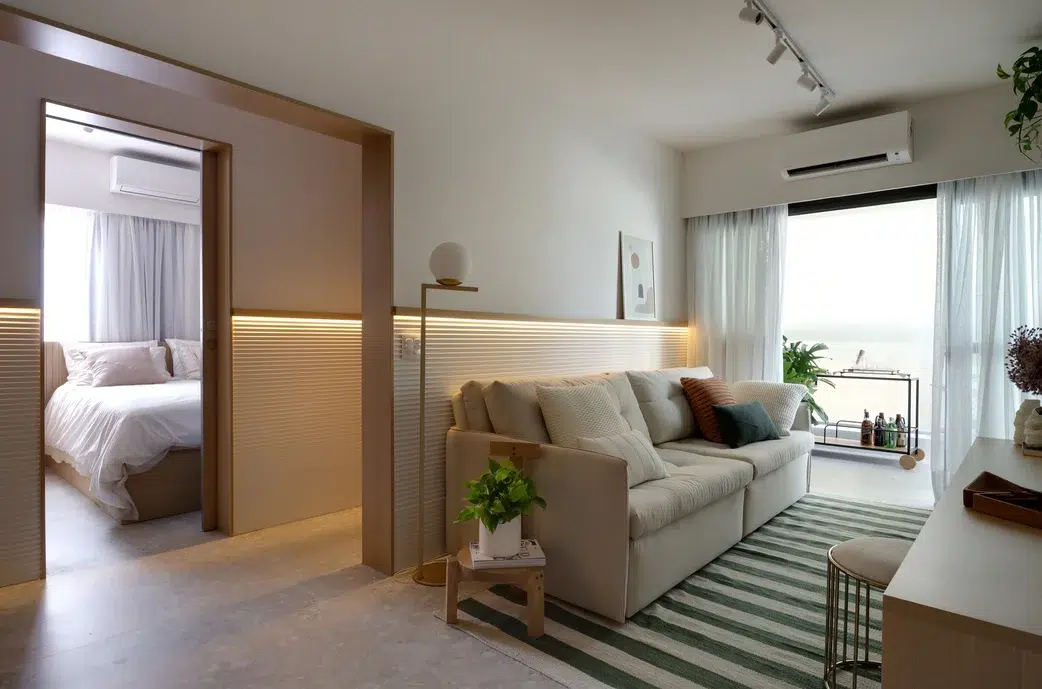 Sala de um apartamento com um sofá off white de dois lugares, tapete de linhas verticais, meia parede com iluminação na parte de baixo e revestimento de madeira, sacada com mini bar e decoração com plantas.